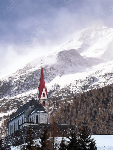 Church with a snowy mountains in the background with wind gusting on slopes and top, Trentino-Alto Adige, Italy photo