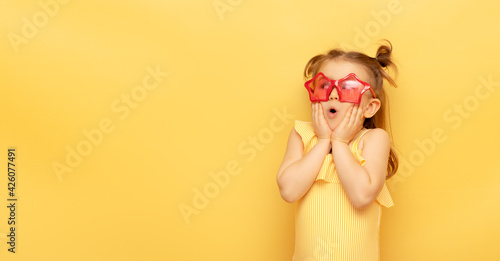 Little child girl in striped swimsuit and red funny summer sunglasses surprised expression looks at camera posing on yellow background, studio portrait.Advertising of children's products and sale