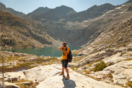 A hiker takes photos of the natural landscape with a reflex camera, in a lake with turquoise waters that reflect the mountains in summer, Aragonese Pyrenees, Huesca, Posets-Maladeta Natural Park.