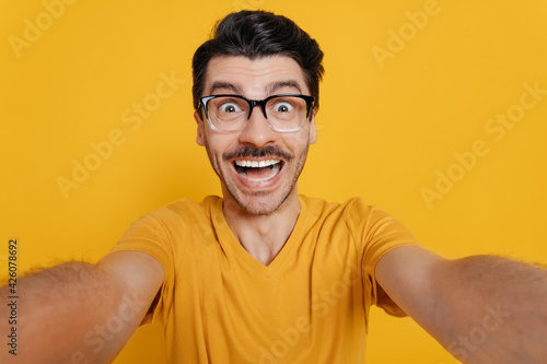 Amazed caucasian unshaved excited guy with crazy gladden face expression, in orange t-shirt and sunglasses, doing selfie shot and smiles into the camera, stands on isolated orange background