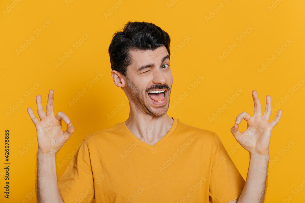 Caucasian unshaved excited young man with happy gladden face expression, in orange t-shirt, shows OK gesture, winks and smiles into the camera, stands on isolated orange background Caucasian unshaved excited young man with happy gladden face expression, in orange t-shirt, shows OK gesture, winks and smiles into the camera, stands on isolated orange background