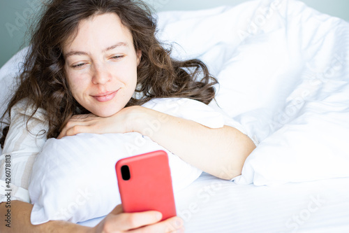 Cuadro en lienzo Beautiful brunette young woman looking at smartphone lying in white bed after sleep in morning, using wireless internet connection at home
