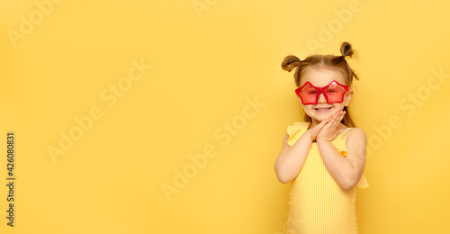 Little child girl in striped swimsuit and red funny summer sunglasses looks at camera posing on yellow background  studio portrait.Advertising of children s products and summer sale. Mockup copy space