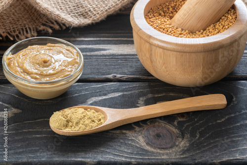 Mustard seeds in a mortar, ground mustard seeds in the spoon and mustard dip in the bowl on brown wooden table