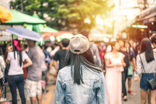 Back of women black hair with jeans jacket and hat on blur crowd anonymous background. She walking and shopping at weekend market. travel concept.