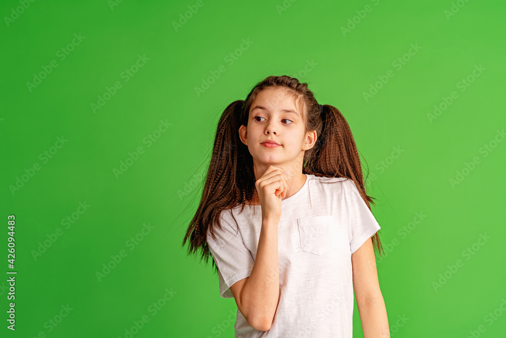 pensive 10-year-old girl in a white T-shirt stands and thinks on a green background. The child ponders his decision, reflects on the correct answer. copy space pensive 10-year-old girl in a white T-shirt stands and thinks on a green background. The child ponders his decision, reflects on the correct answer. copy space