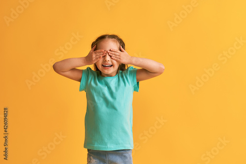 little cute smiling girl covers her eyes with her hands, on a yellow background. child is waiting for a surprise, anticipation and surprise. Copy the space.