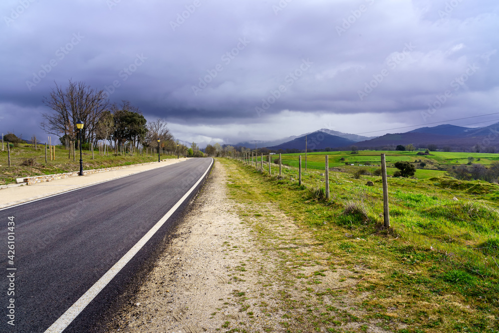 Asphalt road leading to mountains with dark storm cloud sky.