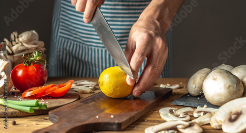 Chef in a blue apron is cutting a lemon with a knife to prepare a delicious dish. Concept of the cooking process