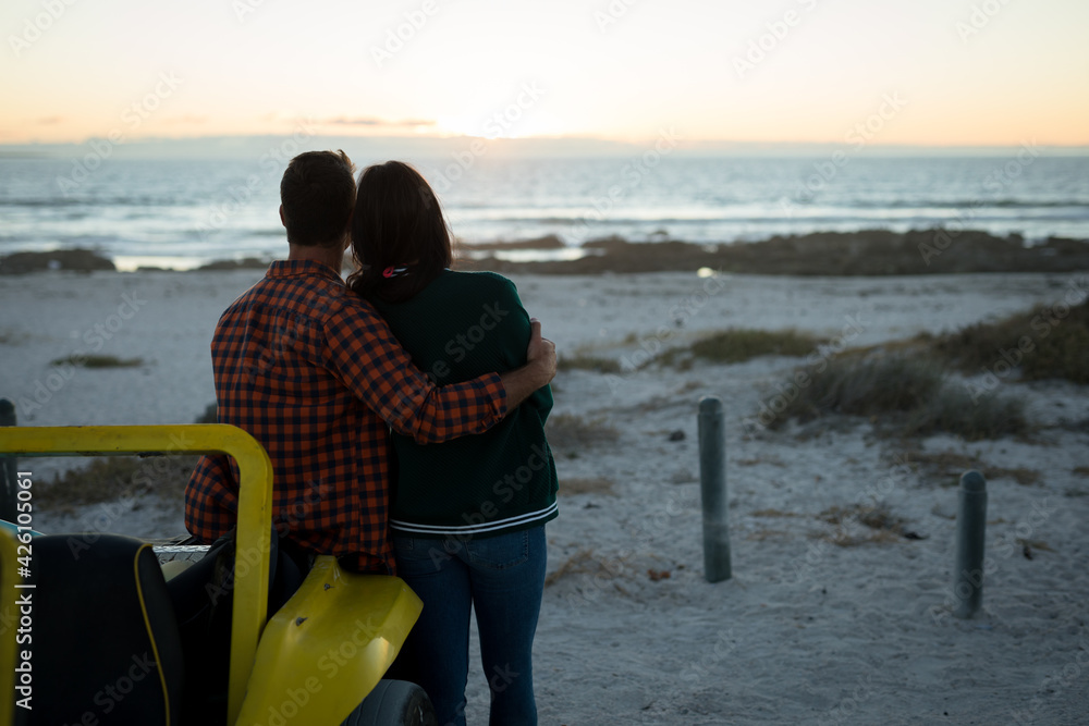 Happy caucasian couple leaning against beach buggy by the sea during sunset embracing - obrazy, fototapety, plakaty 