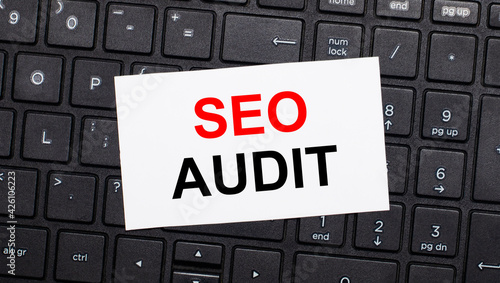 On a black computer keyboard there is a white card with the text SEO AUDIT. View from above