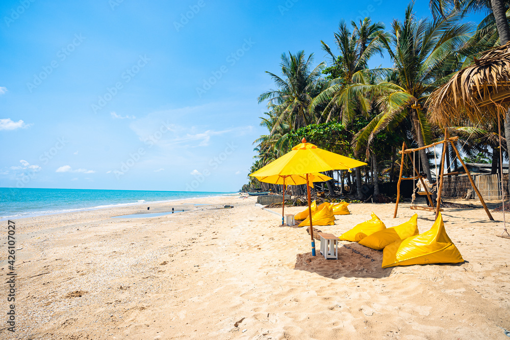 Wooden swing on the beach with yellow umbrella and yellow beach chairs with brown sand, blue sea water, clear blue sky and coconut trees in background.