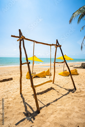 Wooden swing on the beach with yellow umbrella and yellow beach chairs with brown sand, blue sea water, clear blue sky and coconut trees in background.