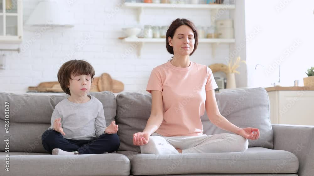 Mum practicing, teaching child to meditate. Caucasian fit mom with cute little son doing yoga exercise sit together on sofa at home. Calm mother and kid sitting in lotus pose on couch. Stress relieve Mum practicing, teaching child to meditate. Caucasian fit mom with cute little son doing yoga exercise sit together on sofa at home. Calm mother and kid sitting in lotus pose on couch. Stress relieve