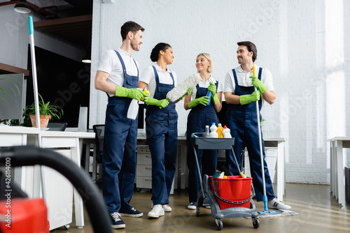 Smiling multiethnic cleaners talking near cart with detergents in office