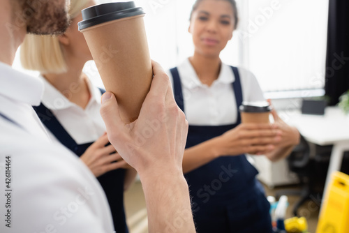 Cleaner holding takeaway drink near interracial workers on blurred background in office