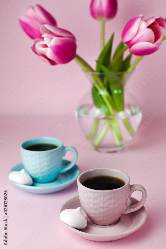 A two cups of coffee and a glass vase with pink tulips on the pink background. Close-up.