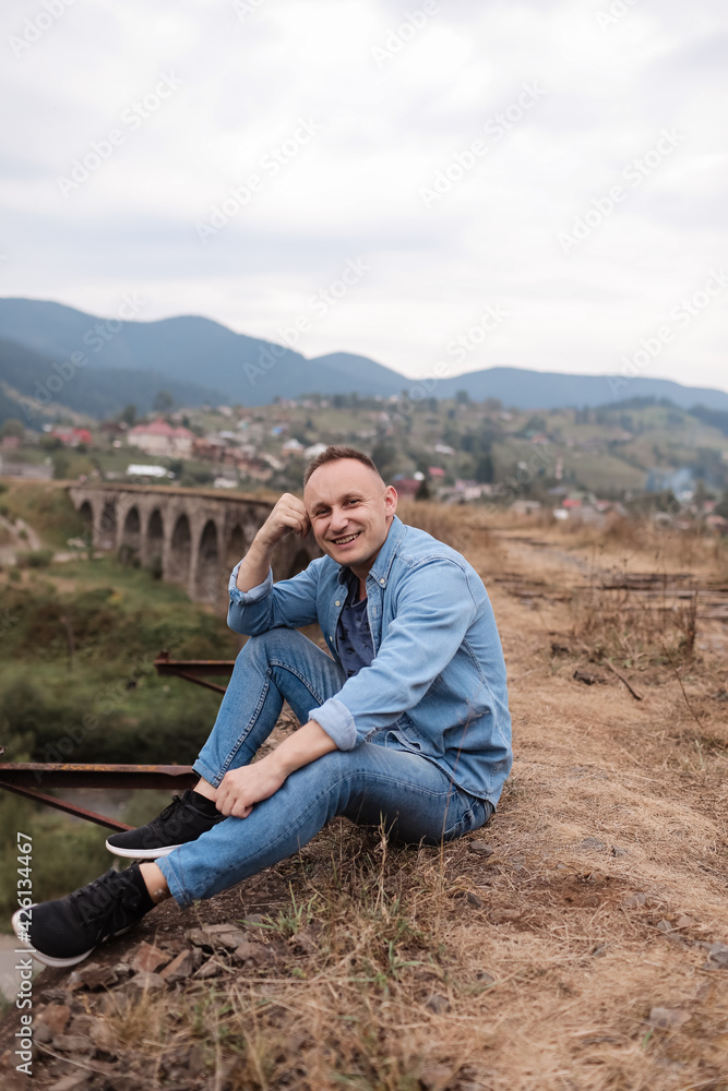Stylish young man on the railway bridge in Vorokhta, Carpathians against the backdrop of a mountain landscape. tourists sitting in the mountains near the railway