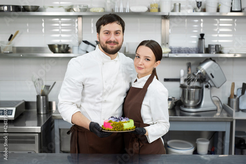 Happy loving couple of pastry chefs, a man and a woman hold a mousse cake in their hands in a professional kitchen.