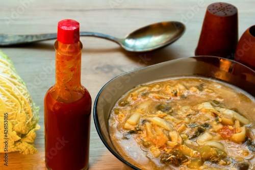 Spicy asian noodle soup in a glass bowl, tabasco sauce and chinese cabbage served on a wooden board
