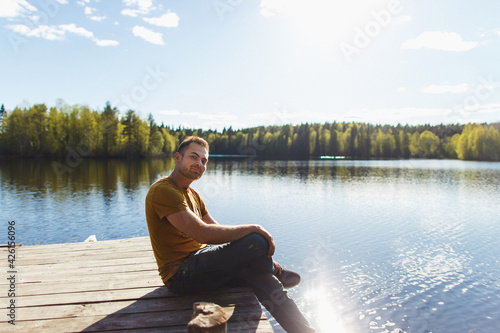 Young handsome man sitting near forest lake, relaxing and meditating in spring or summer sunny day