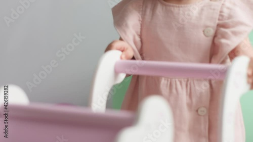 baby girl in a pink dress at home walks with baby wooden stroller