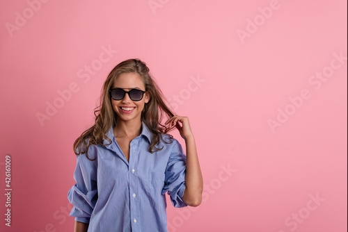 a happy young girl in a blue shirt and sunglasses on a pink background. the concept of summer, sunny weather, joy and fun © Асель Иржанова