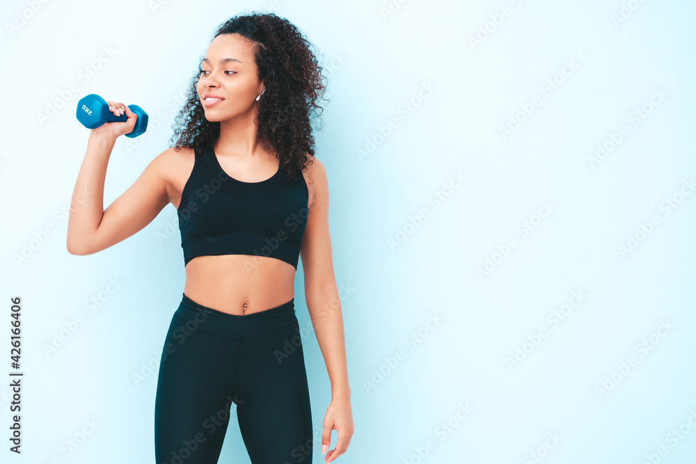 Fitness smiling black woman in sports clothing with afro curls hairstyle.She wearing sportswear. Young beautiful model with perfect tanned body.Female holding dumbbells in studio near light blue wall Fitness smiling black woman in sports clothing with afro curls hairstyle.She wearing sportswear. Young beautiful model with perfect tanned body.Female holding dumbbells in studio near light blue wall
