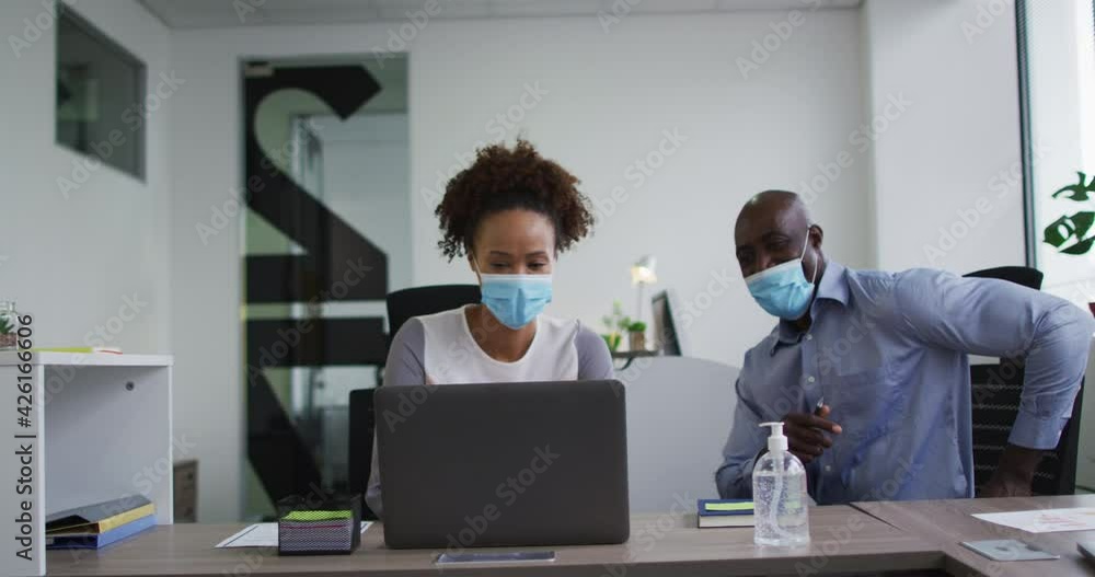Diverse businessman and businesswoman in face masks discussing, using laptop in office Diverse businessman and businesswoman in face masks discussing, using laptop in office