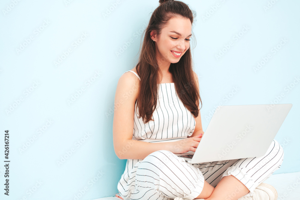 Young beautiful smiling female in trendy summer hipster overalls clothes. Sexy carefree woman posing near light blue wall in studio. Positive model having fun indoors.Using her computer laptop Young beautiful smiling female in trendy summer hipster overalls clothes. Sexy carefree woman posing near light blue wall in studio. Positive model having fun indoors.Using her computer laptop