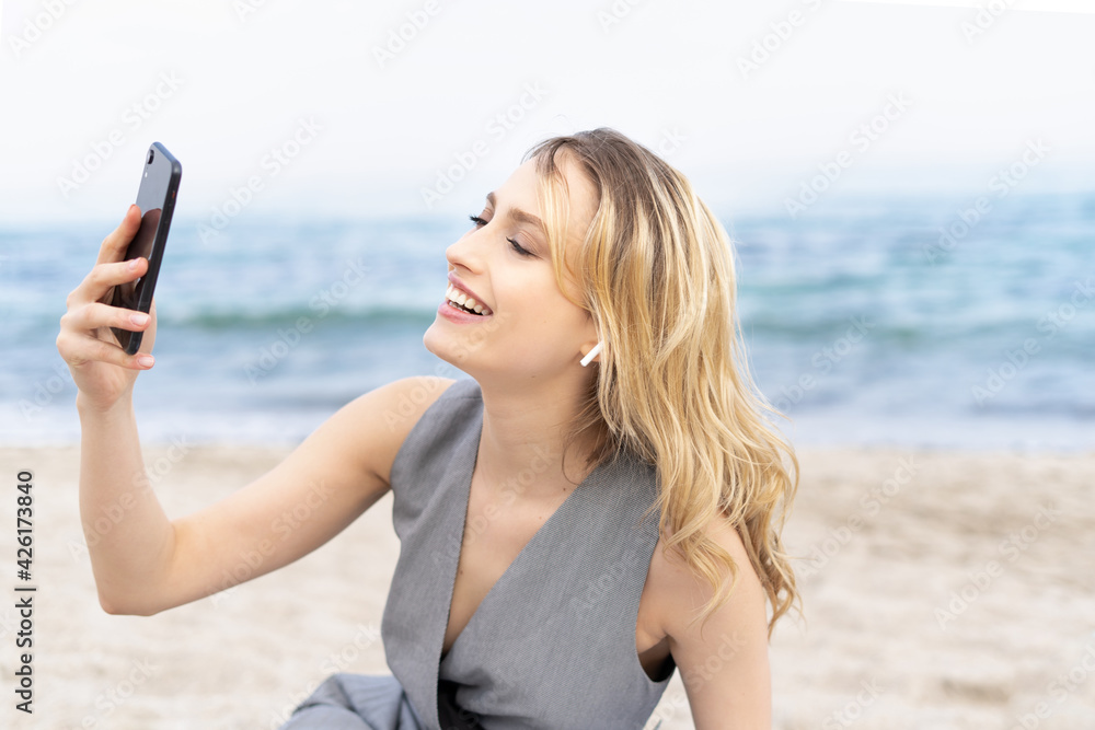 Cheerful blonde girl laughing through the video call on her mobile phone sitting on a sand near the sea 