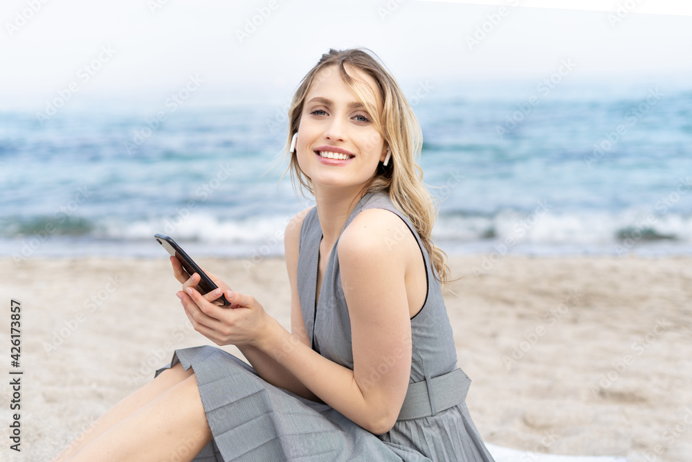 Charming young blonde women smiling holding a mobile phone and wearing a headphones sitting on a sand beach near the sea 