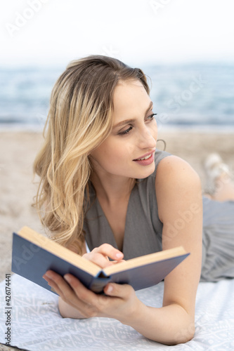 Close up of a cheerful young blonde woman with a book lies on the beach and looking right 