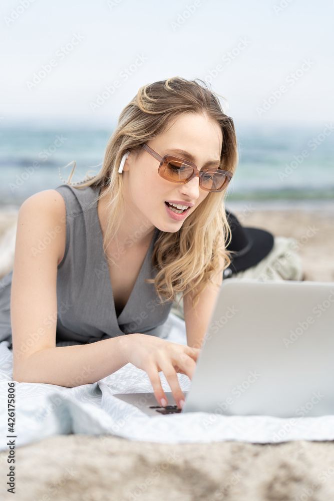 Stylish active young woman working remotely on the laptop computer wearing sun glasses and wireless headphones, talking on a zoom video chat lying on a beach  
