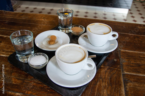 Two cups of coffee latte on a rustic wooden table with sugar and cup of water and a small cookie in Rome. 