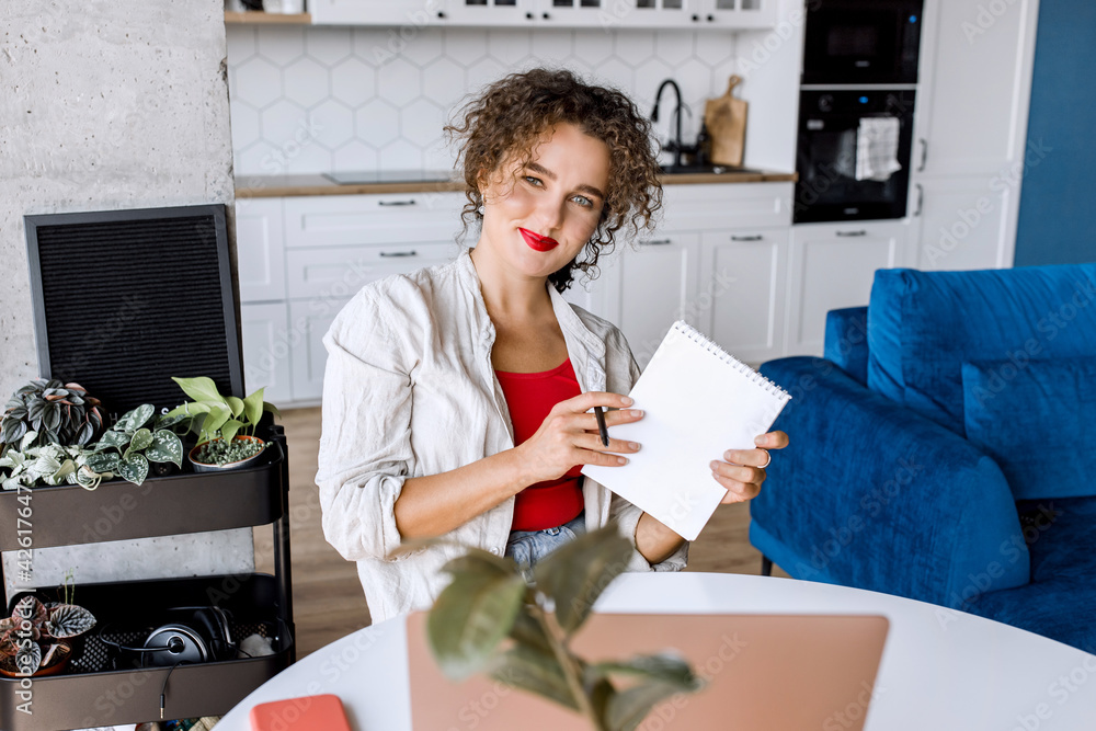 Portrait of confident stylish business woman is making notes, looking at the camera, smiling. Successful young female freelancer or student remotely working or studying at home Portrait of confident stylish business woman is making notes, looking at the camera, smiling. Successful young female freelancer or student remotely working or studying at home