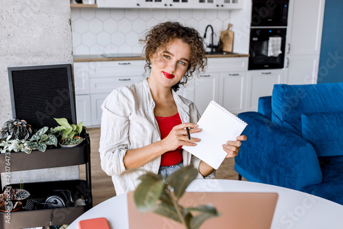 Portrait of confident stylish business woman is making notes  looking at the camera  smiling. Successful young female freelancer or student remotely working or studying at home