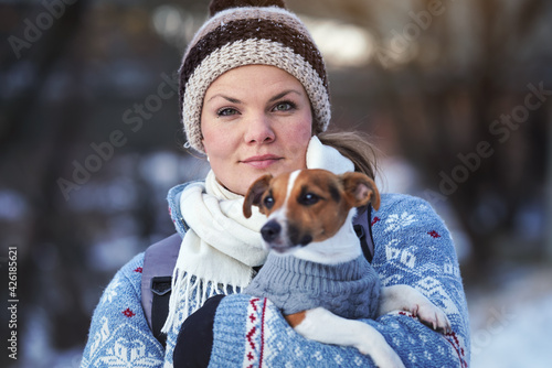 Young woman in winter jacket holding her Jack Russell terrier dog on hands, blurred trees background