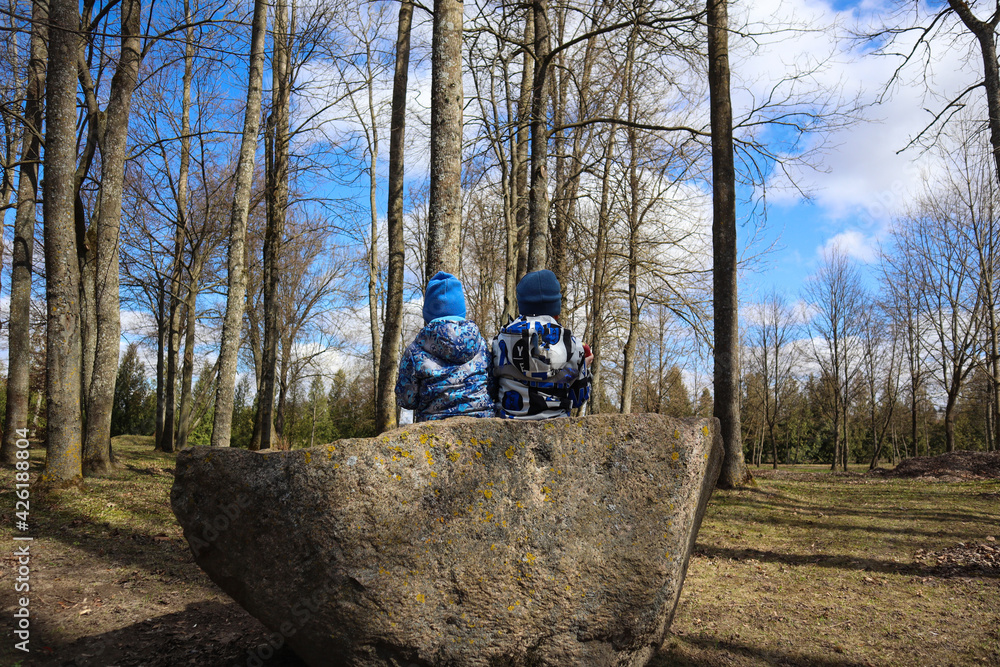 Obraz premium Two children in blue jackets sit on a large boulder in an old park on a spring day, side view