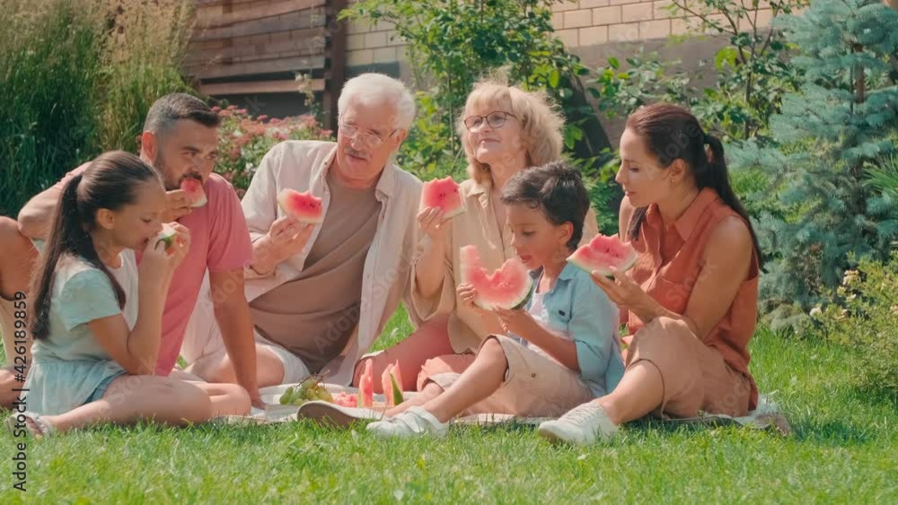 Full PAN shot of delighted multigenerational family of six spending summer day together sitting on grass in backyard chatting and eating watermelon Full PAN shot of delighted multigenerational family of six spending summer day together sitting on grass in backyard chatting and eating watermelon