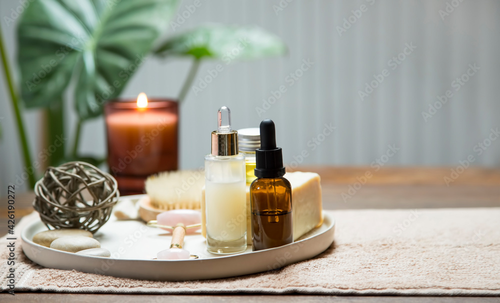 clean organic skincare and bodycare products with oil bottle, serum, natural soap and quartz roller with candle and green plants in the background