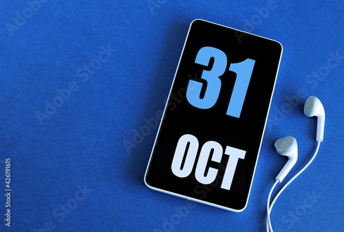 October 31. 31 st day of the month, calendar date. Smartphone and white headphones on a blue background. Place for your text. Autumn month, day of the year concept