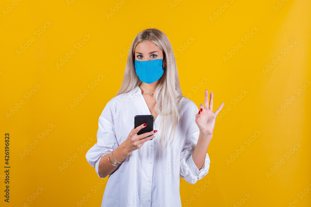 Woman wearing face mask protect from virus showing ok sign. Young girl wearing face light blue mask using smartphone and showing ok sign, isolated over yellow background Woman wearing face mask protect from virus showing ok sign. Young girl wearing face light blue mask using smartphone and showing ok sign, isolated over yellow background