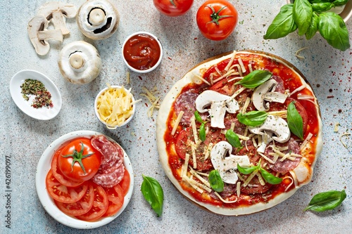 Cooking Italian pizza with tomato sauce, fresh tomatoes, cheese, mushrooms, salami slices and basil . Fresh food cooking concept at home. Semi-finished product, raw dough.