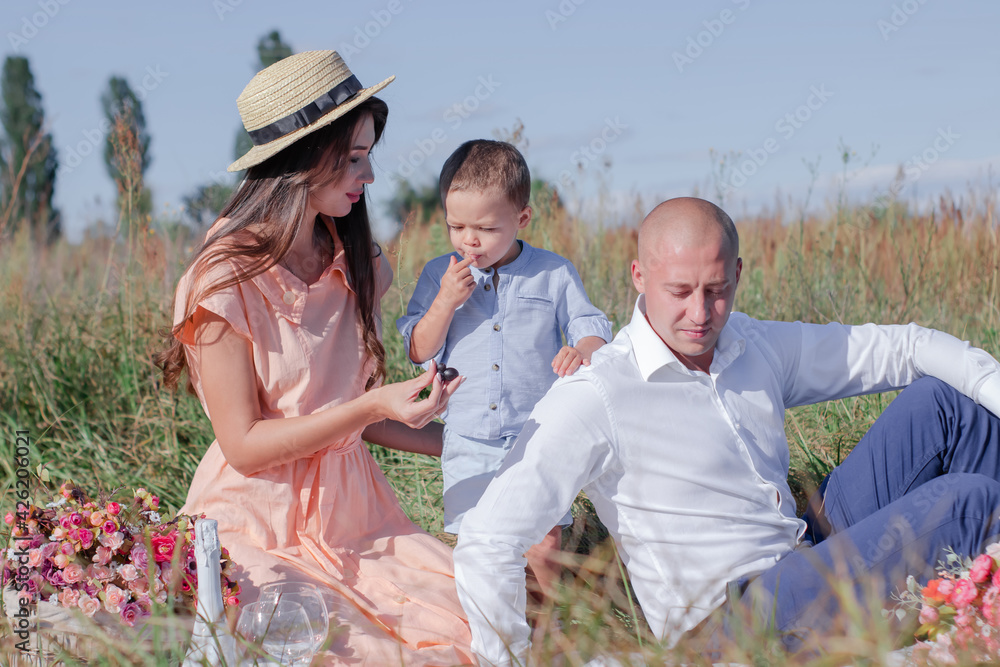 happy family on a picnic in the field. husband, wife and little boy sitting on a blanket with flowers, wine, baguette and fruits nearby. romantic date. parenting. mother, father and son together