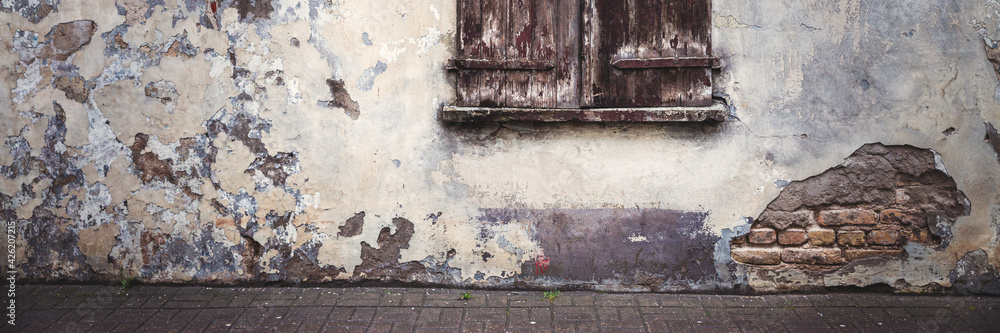 Naklejka premium Old wall with peeling plaster. Window covered with wooden shutters. Weathered rough surface of the wall. Medieval European city. Vintage texture for background and design.