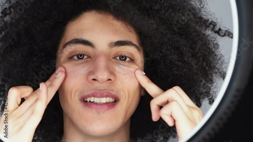 Man skincare. Facial treatment. Anti aging cosmetic product. Confident handsome guy with long curly hair applying smearing face cream under eyes in round mirror reflection.