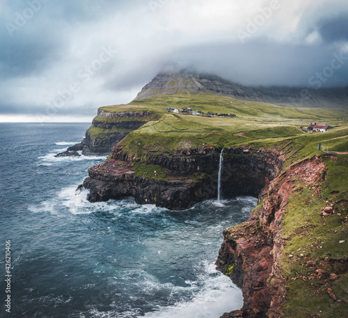 Aerial drone view of Gasadalur village and Mulafossur its iconic waterfall, Vagar, Faroe Islands, Denmark. Rough see in the north atlantic ocean. Lush greens during summer.