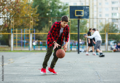 Cute young boy plays basketball on street playground. Teenager in red  flannel checked shirt with orange basketball ball outside. Hobby, active lifestyle, sport activity for kids. © Natali