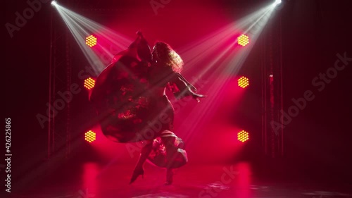 Silhouette of a passionate young woman in a gypsy dress whirls dancing and waving a long skirt. Female dancer dancing romany folk dance in dark smoky studio with red light. Slow motion.